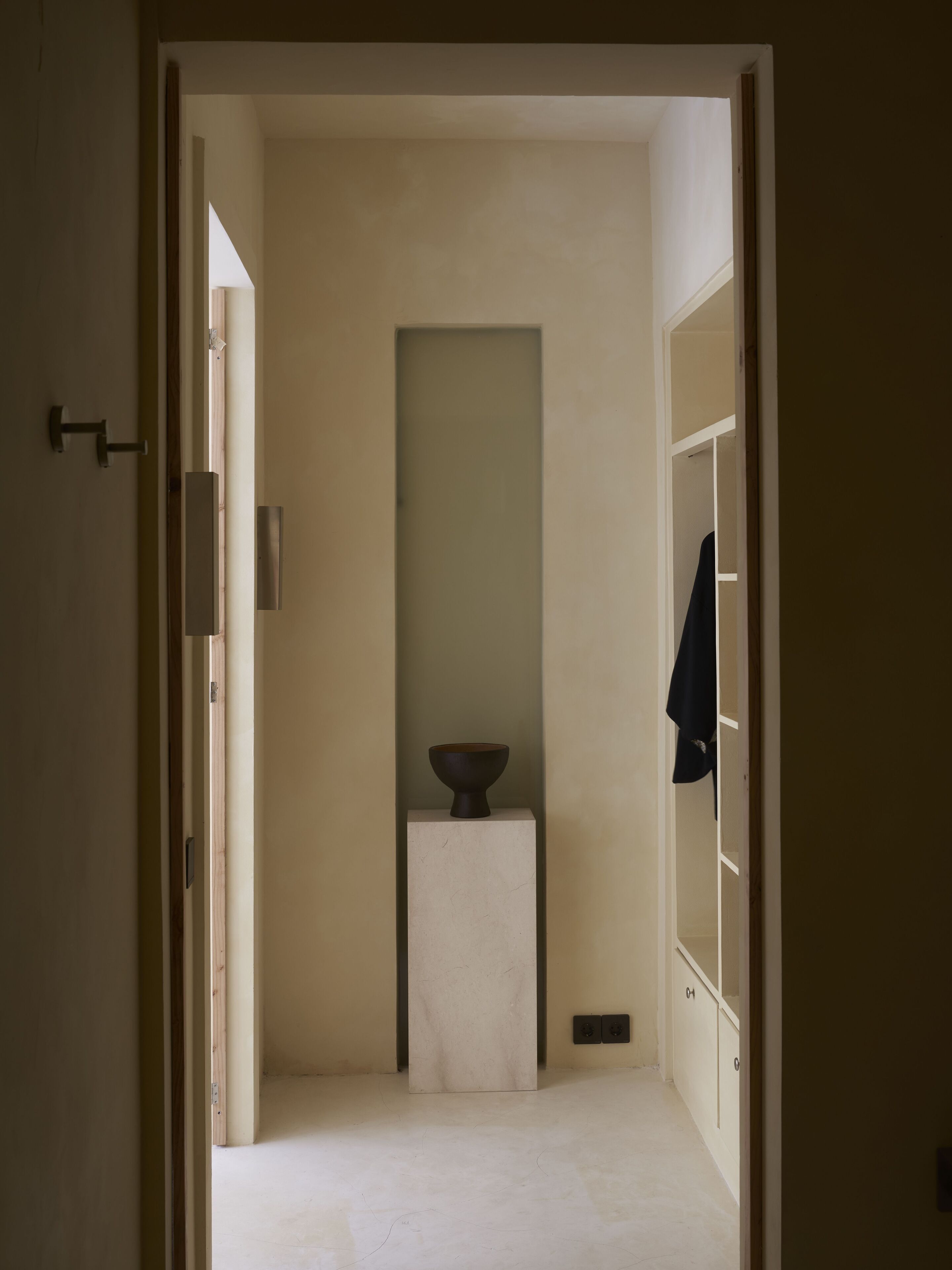 A narrow hallway with a small table and a bowl in the center, leading to a closet with hanging clothes.