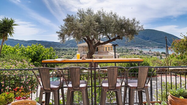 Une terrasse avec une table et des chaises, donnant sur un paysage montagneux et offrant une vue sur une étendue d’eau et des bâtiments.