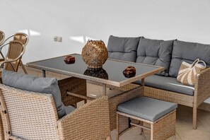 Un canapé en osier avec des coussins gris, une table en osier avec un plateau en verre et un vase tressé.