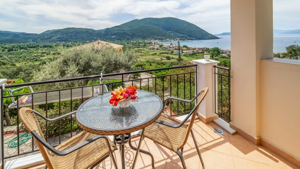 A balcony with wicker furniture, a glass table with a vase of flowers, and a view of green hills and a body of water. - Studio Apartment 'Panorama 1' with Sea View, Wi-Fi and Air Conditioning (Lefkada)