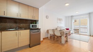 Una cocina moderna con armarios de madera, lavavajillas y microondas. Hay un comedor con mesa y sillas, y un balcón con vistas.
