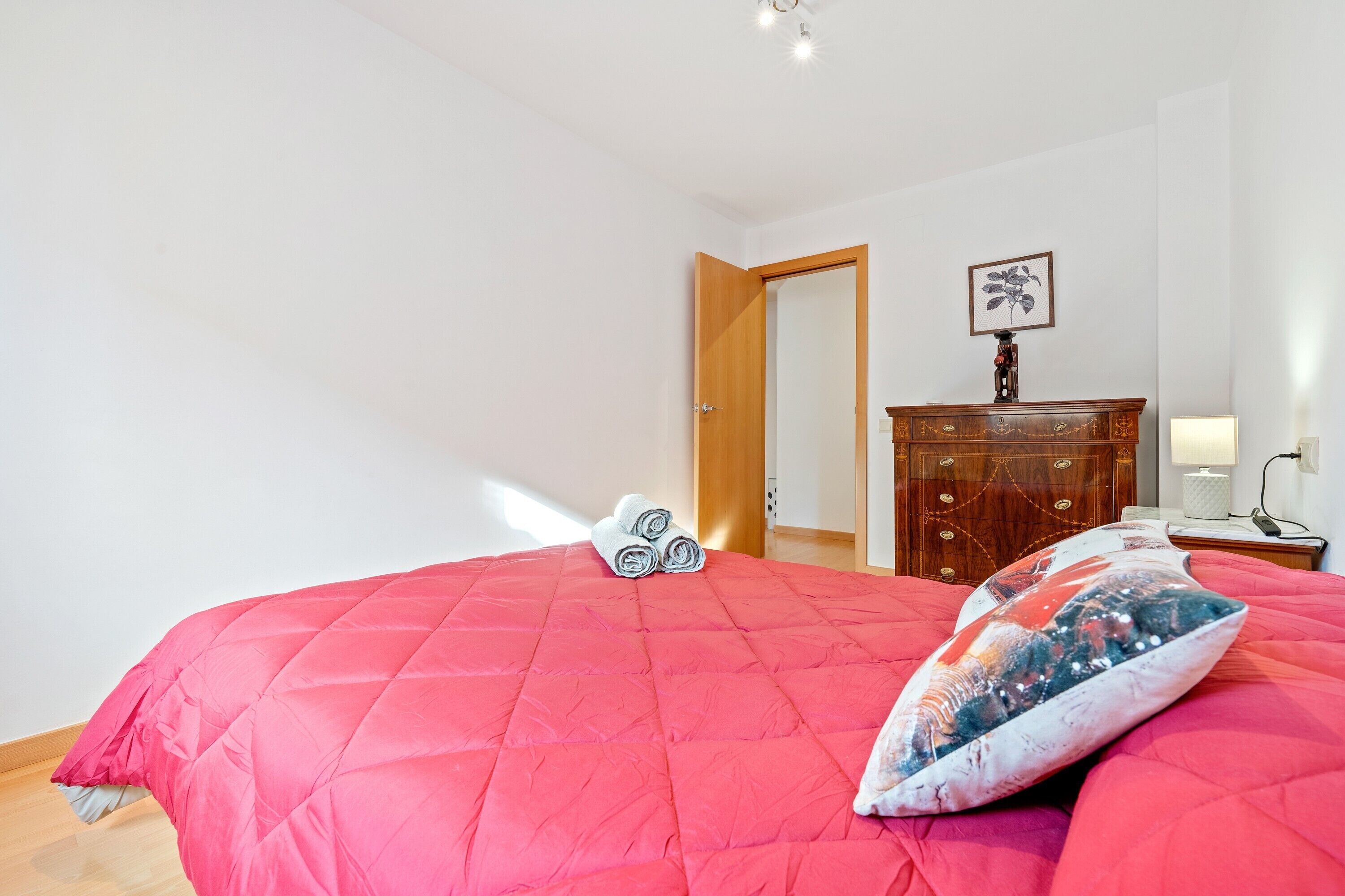Une chambre à coucher avec une couvre-lit rouge, une commode en bois et une porte donnant sur une autre pièce.