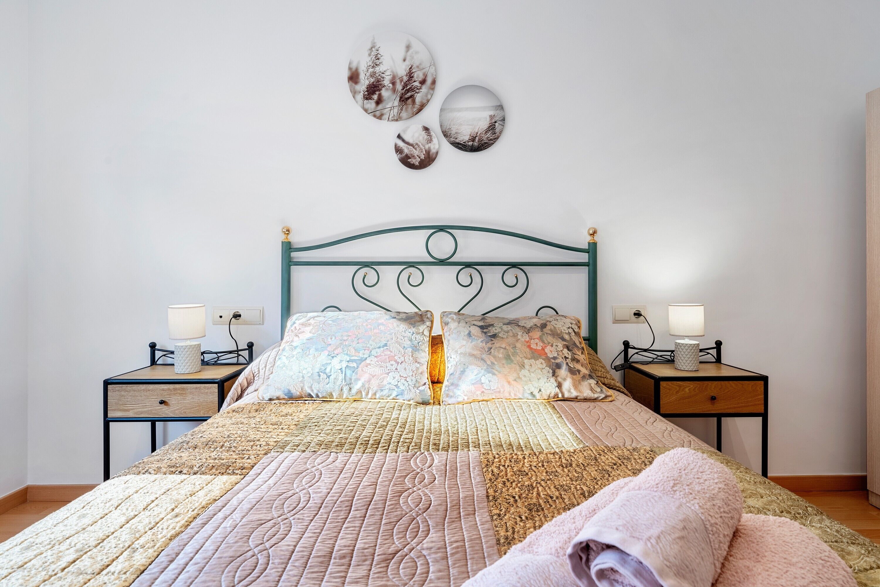 Una cama bien hecha con colcha, dos lámparas de noche y una pared decorada con tres piezas de arte circular.