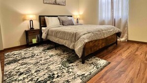 A bedroom with a bed, a nightstand, a lamp, a painting, and a window with curtains.