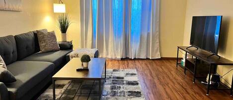 A living room with a sofa, coffee table, TV stand, and a large window with curtains.