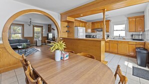 Une cuisine moderne avec des meubles en bois, un coin repas comprenant une table et des chaises en bois, et une vue sur un salon avec un canapé et une table basse, accessible par un grand arc.