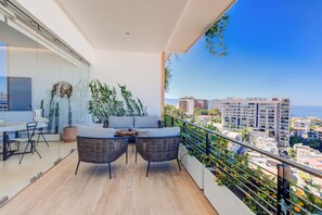 Un balcone con mobili in vimini, un tavolo in vetro e vista sulla città e sul mare.