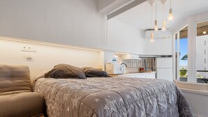 Quarto com cama, sofá, janela com vista e uma cozinha compacta ao fundo.