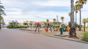 Ein gepflasterter Weg, Spielgeräte, Palmen und Gebäude im Hintergrund.