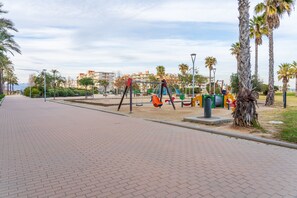 A paved walkway, playground equipment, palm trees, and buildings in the background. - Apartment 'Tirreno 304' with Sea Views, Private Terrace and Wi-Fi (La Pineda)
