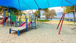 A playground with a slide and climbing structures, located on sandy ground with trees and a body of water in the background. - Causeway's Pinnacle! (Yeppoon)