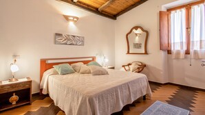 Une chambre avec un plafond en bois, un lit recouvert d’une courtepointe blanche, deux lampes de chevet, une table de chevet avec une lampe, une chaise et une fenêtre avec des rideaux.