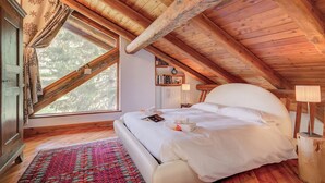 A cozy attic bedroom with a large bed, wooden beams, a window with curtains, and a small bedside table with a lamp.