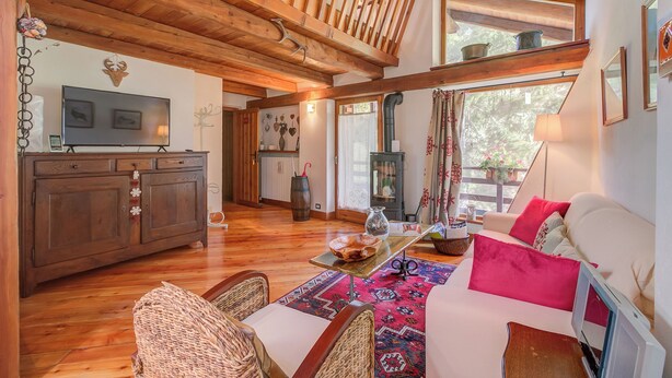 A cozy living room with a wooden ceiling, a sofa with red cushions, a television, and a dining area with a table and chairs.