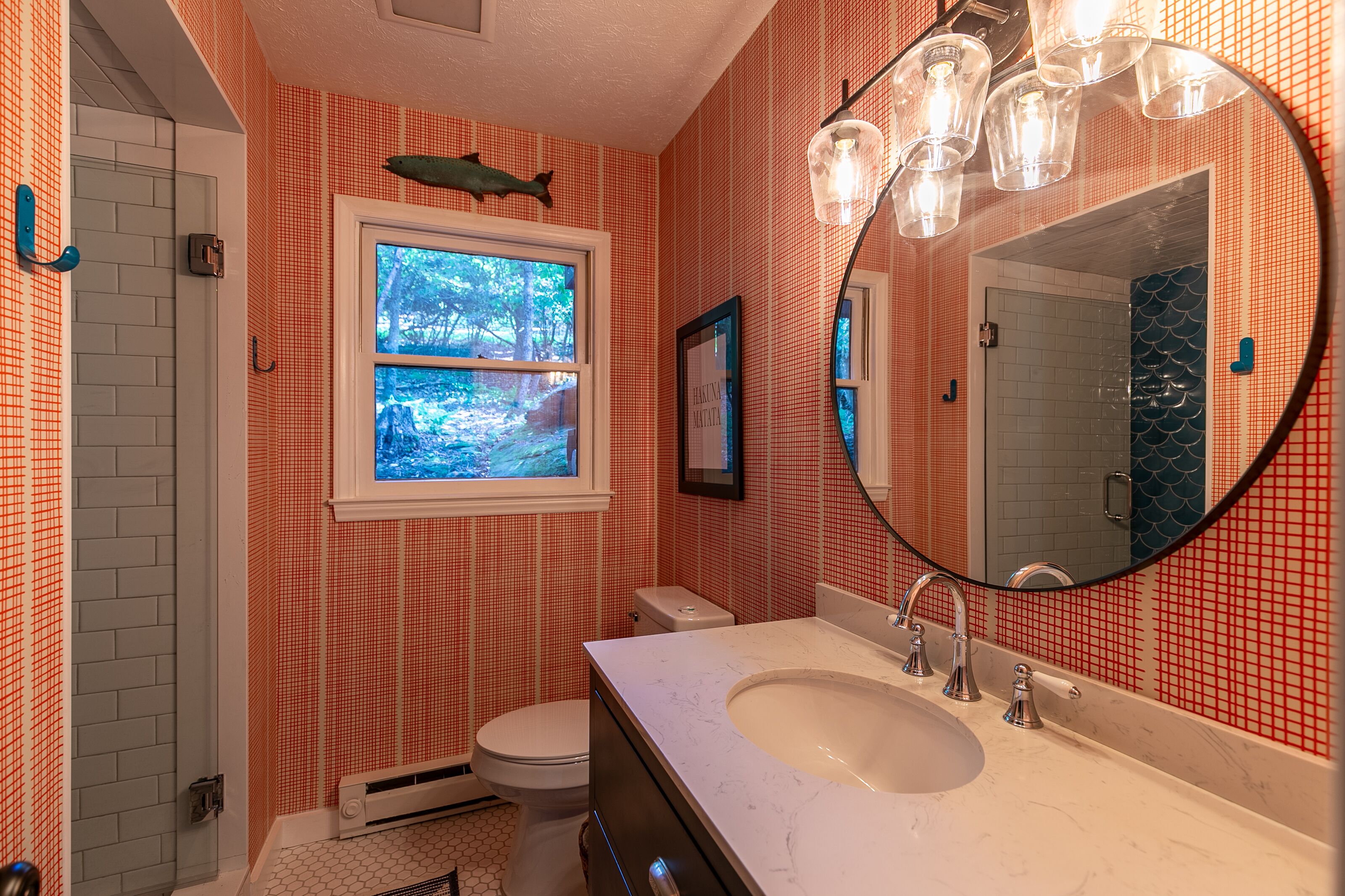 Une salle de bain avec un lavabo blanc, un grand miroir et une fenêtre donnant sur des arbres.