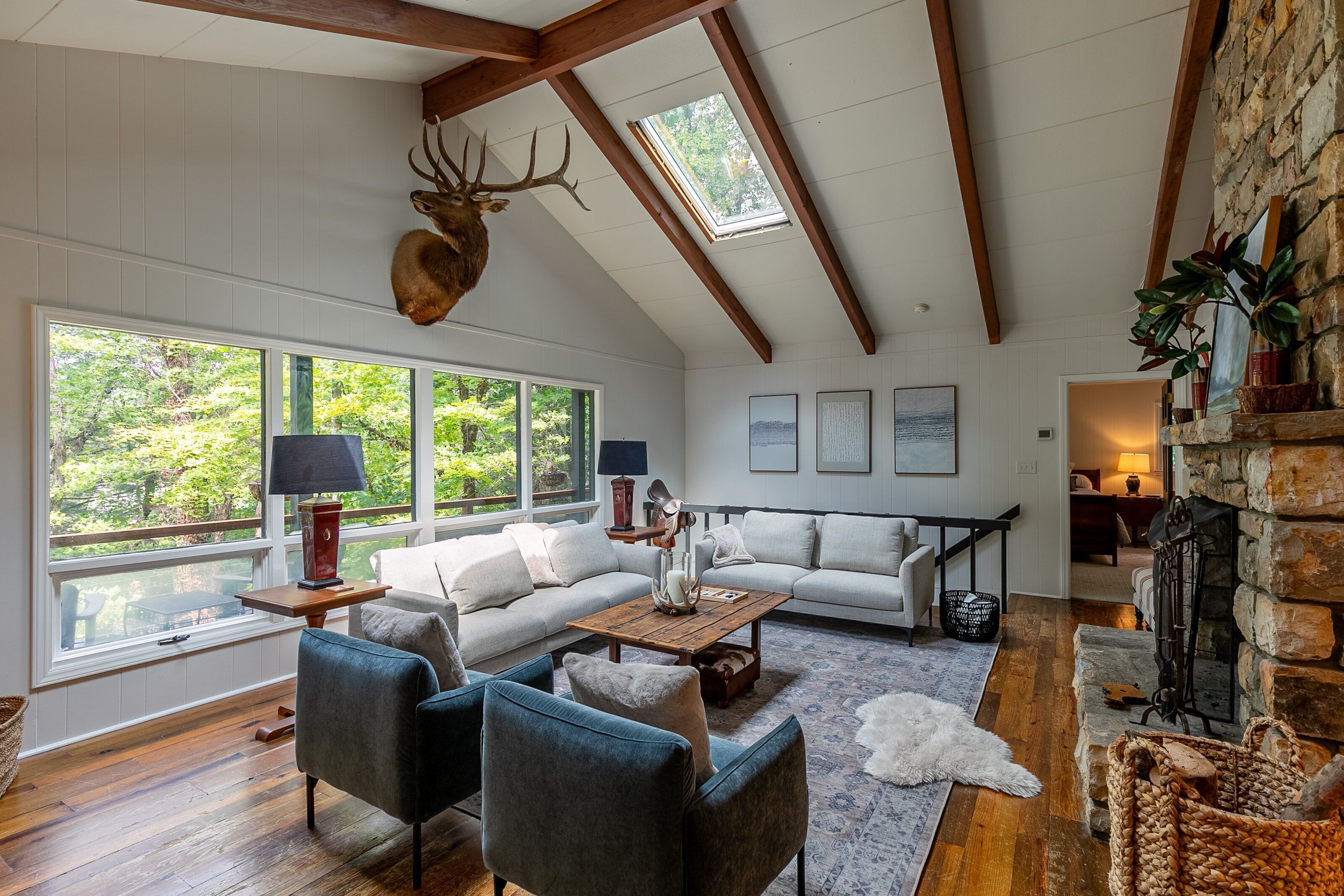 A spacious living room with a high ceiling, a stone fireplace, and a large window overlooking a forest.