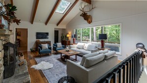 Ein geräumiges Wohnzimmer mit hoher Decke, Holzbalken, einem Steincheminée und einem aufgehängten Hirschkopf.
