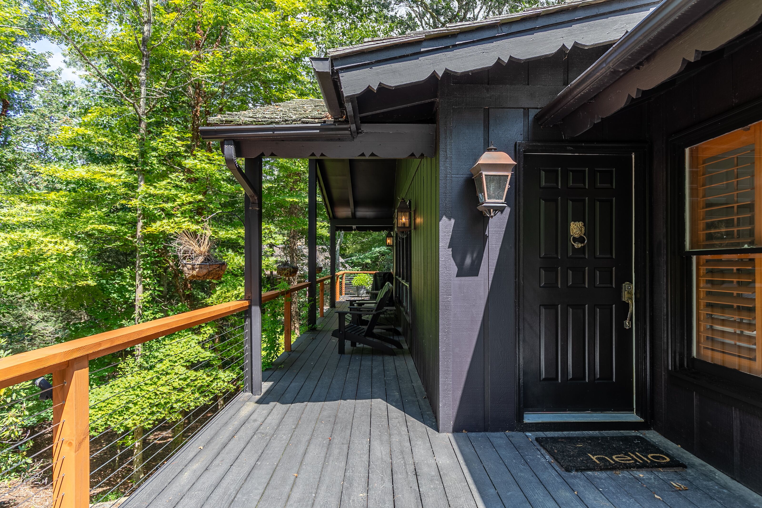 Une terrasse en bois avec une porte noire et un paillasson sur lequel est écrit « bonjour », le tout entouré d’une végétation luxuriante.
