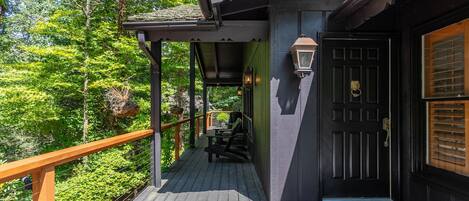 Une terrasse en bois avec une porte noire et un paillasson sur lequel est écrit « bonjour », le tout entouré d’une végétation luxuriante.