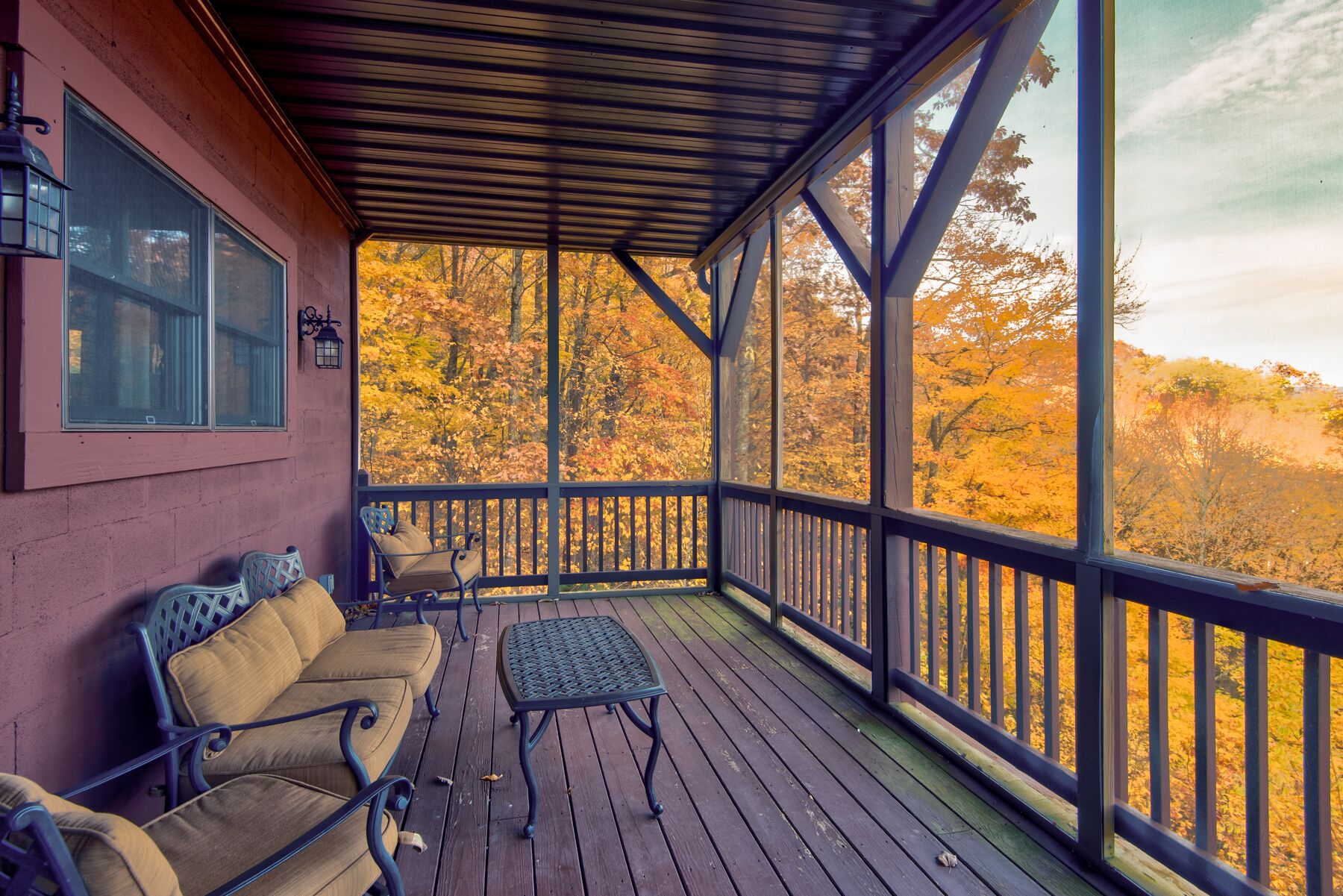 A covered porch with a wooden bench and table, a window, and a view of autumn trees.