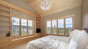 A bedroom with a large bed, wooden shelving, a television, and large windows with a view of trees and mountains.