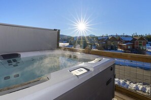 A hot tub with a control panel, surrounded by a wooden deck, overlooking a residential area with houses and a snowy landscape.
