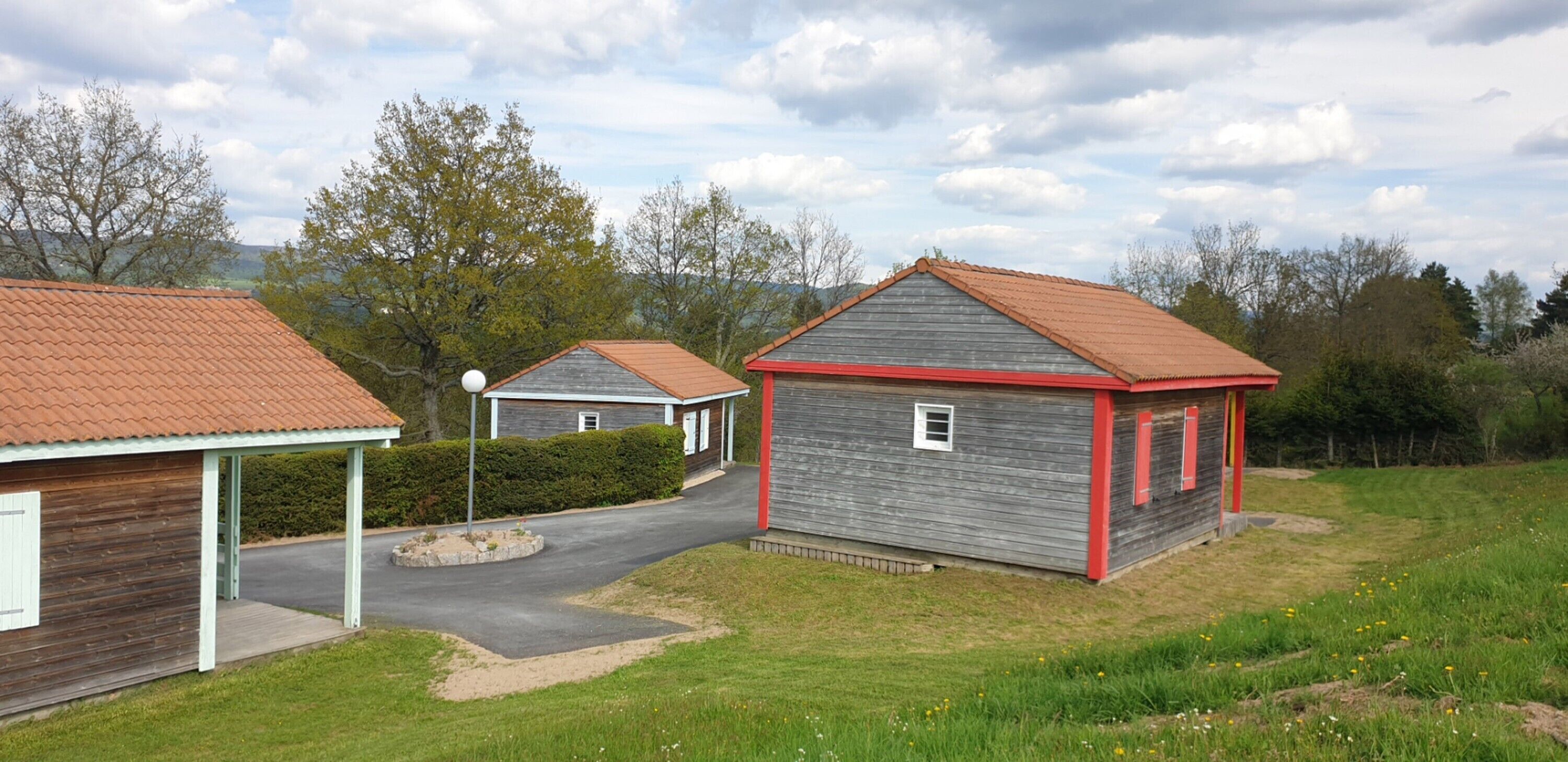 Un paysage rural avec deux bâtiments en bois, l’un avec un toit rouge et l’autre avec un toit gris, entourés de verdure et sous un ciel dégagé.