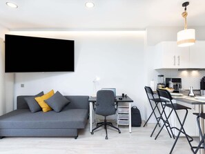 A modern living space with a sofa, desk, and dining area, featuring a flat-screen TV, a lamp, and a kitchen with a coffee machine.
