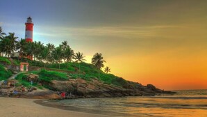 A beach with a lighthouse, palm trees, and rocky terrain. - Amã Stays & Trails Lily Pad , Kumarakom (Kottayam)