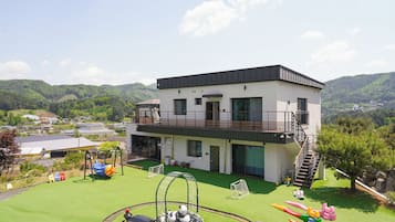 Un edificio de dos pisos con un balcón, una zona de juegos con tobogán y columpios, y vistas a un paisaje montañoso.