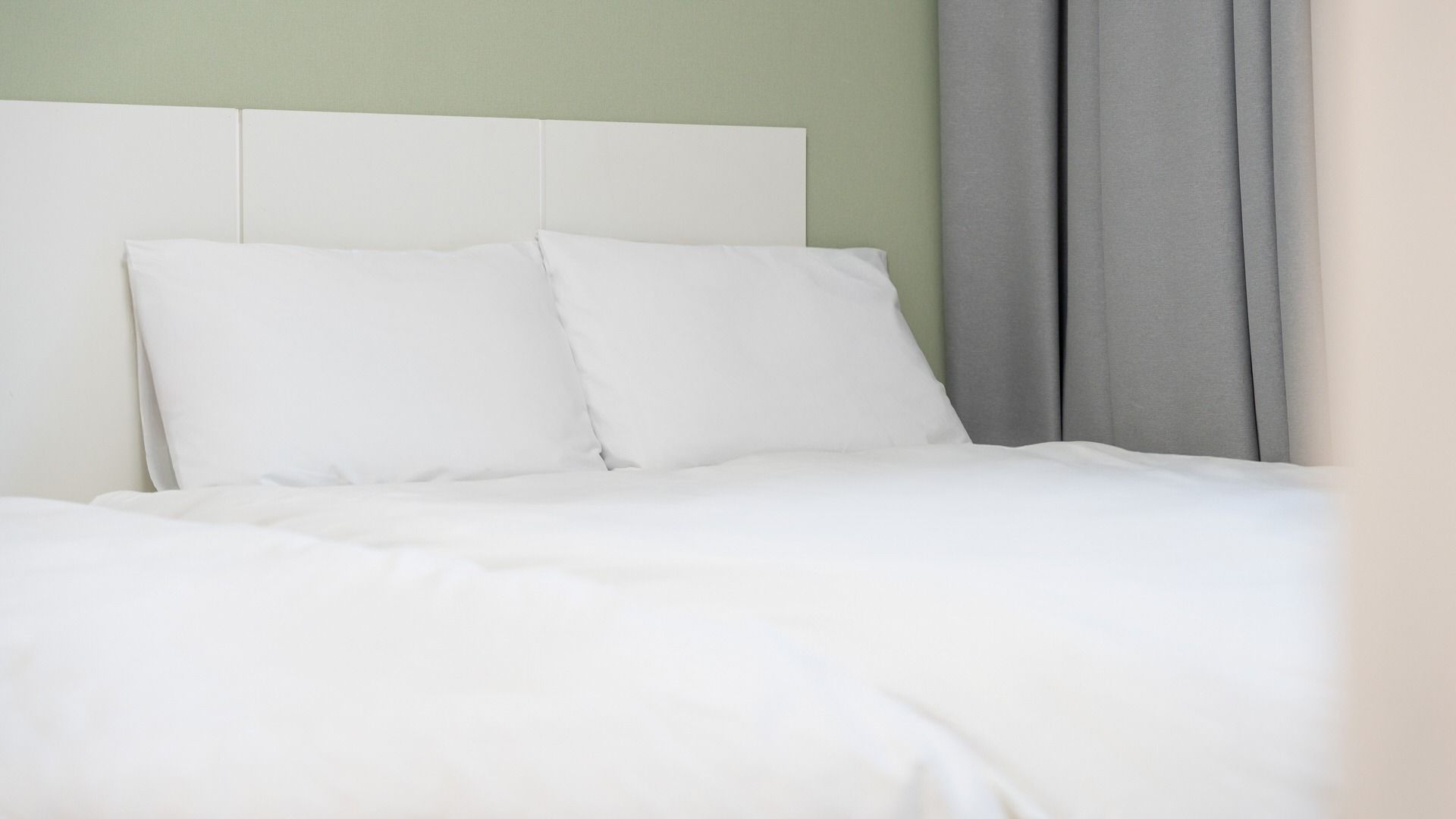 A bed with white bedding and pillows, a white headboard, and a grey curtain.