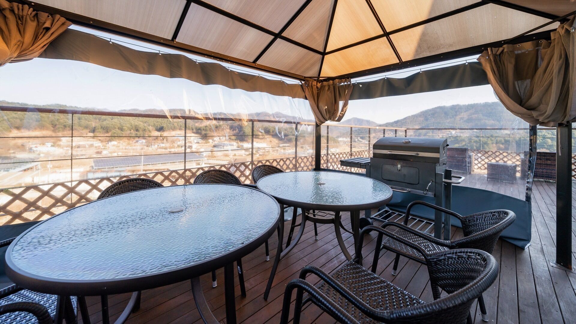Une terrasse extérieure avec des tables rondes et des chaises en osier, un barbecue et une vue sur un paysage vallonné.