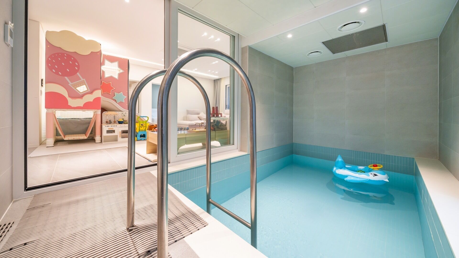 A modern indoor swimming pool with a blue interior, a child-friendly play area with toys and a slide, and a view into a living room with a sofa and a table.