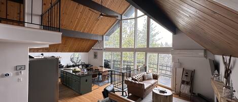 Un loft spacieux avec de hauts plafonds, des poutres apparentes, un coin repas, un espace salon confortable et de grandes fenêtres offrant une vue sur l’extérieur.