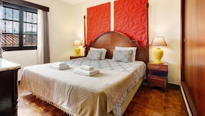 A hotel room with a large bed, two bedside tables with lamps, a window with metal bars, and a red geometric wall decoration.