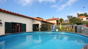 A swimming pool with a tiled roof, a wooden deck, and a building with green shutters.