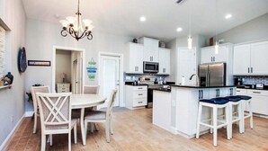 Une cuisine moderne avec des meubles blancs, un îlot central doté de tabourets, un lustre et un coin repas avec une table et des chaises.