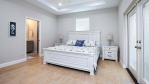Une chambre avec un lit blanc, un ventilateur de plafond, une fenêtre avec des stores, une table de chevet avec une lampe et une porte avec une serrure.