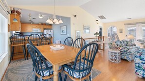 A spacious living area with a wooden dining table, blue upholstered chairs, a patterned sofa, and a kitchen in the background.