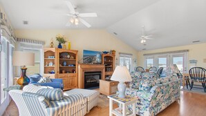 A spacious living room with striped sofas, a floral couch, and a fireplace. There are ceiling fans, a dining area with chairs, and large windows.