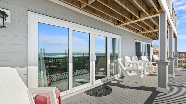 A covered outdoor deck with white chairs and tables, large sliding glass doors, and a view of the ocean.