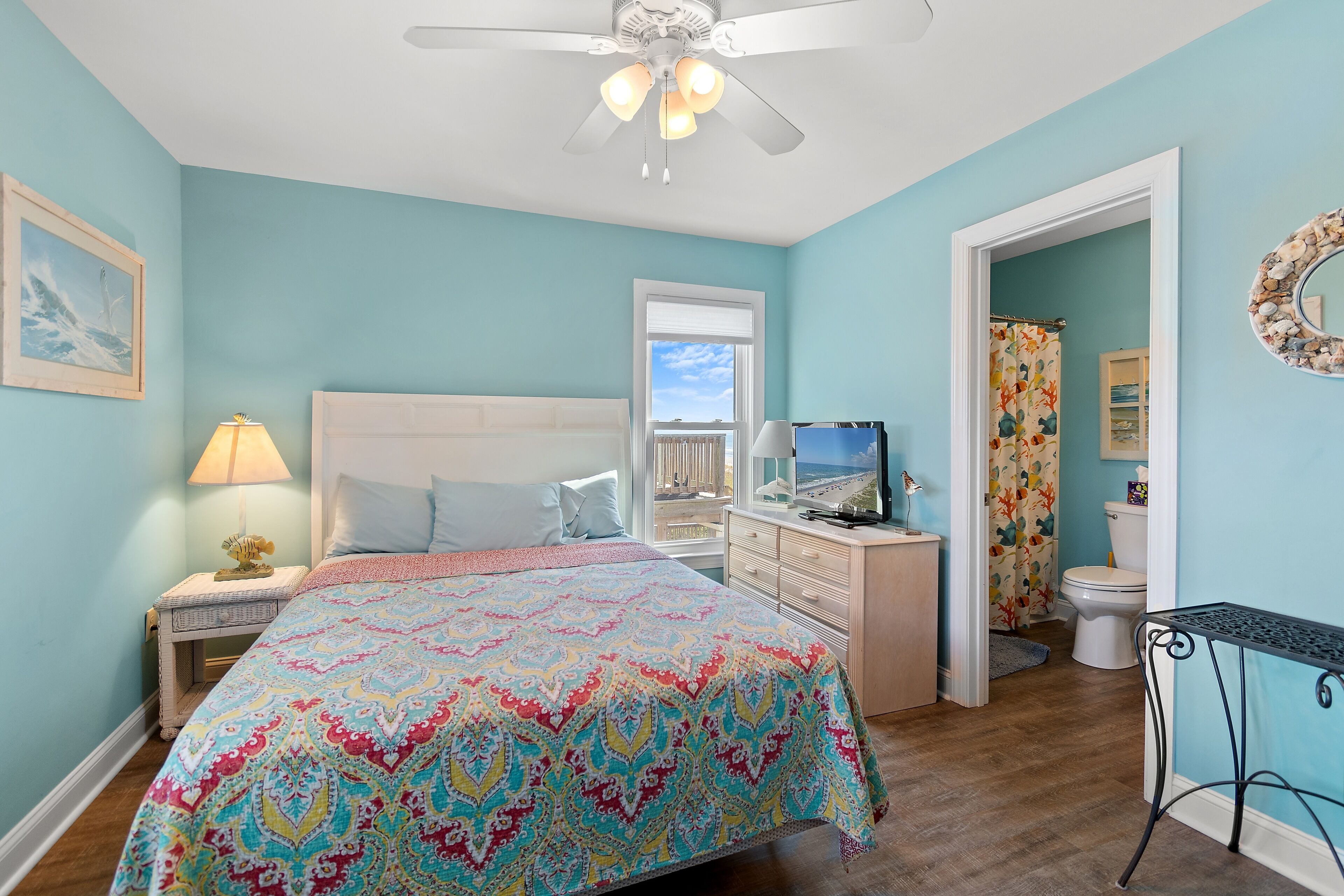 Une chambre à coucher comprenant un lit, une table de chevet, une commode, un ventilateur de plafond, une fenêtre et une salle de bain visible à travers une porte ouverte.