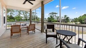 A covered porch with wooden furniture, a ceiling fan, and large windows offering a view of trees and buildings. - "Just Peachy" 3BR Home | 4 Baths (2 Full, 2 Half) (Oak Island)