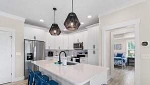 A modern kitchen with white cabinets, a stainless steel refrigerator, a microwave, and a large island with bar stools. - "Just Peachy" 3BR Home | 4 Baths (2 Full, 2 Half) (Oak Island)