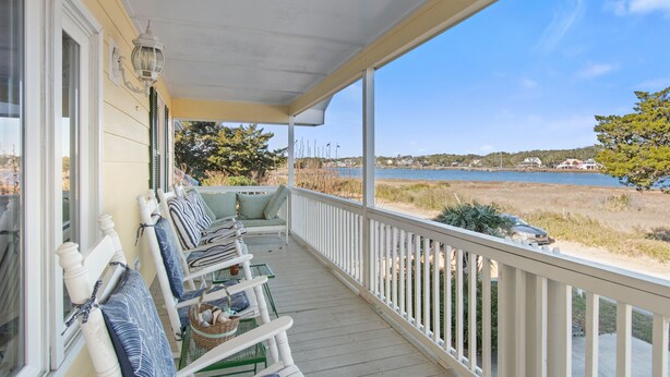 Une véranda couverte avec des balustrades blanches, des meubles d’extérieur verts et une vue sur un lac et une plage.
