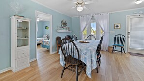 Un coin repas avec une table dressée pour quatre personnes, entourée de chaises en bois. Un ventilateur de plafond est visible au-dessus de la table.