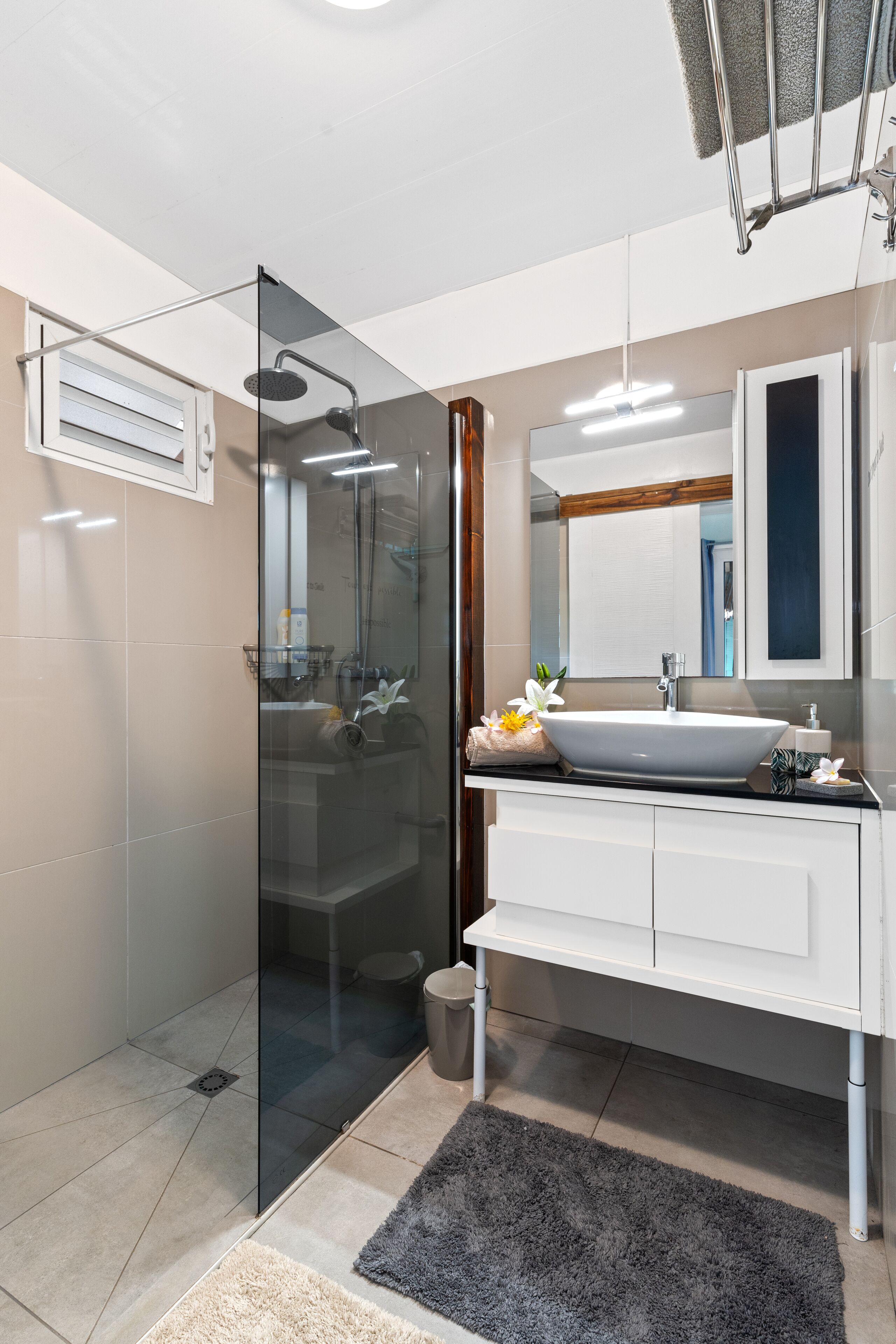 A modern bathroom with a glass-enclosed shower, a white vanity with a large mirror, and a gray rug on the floor.
