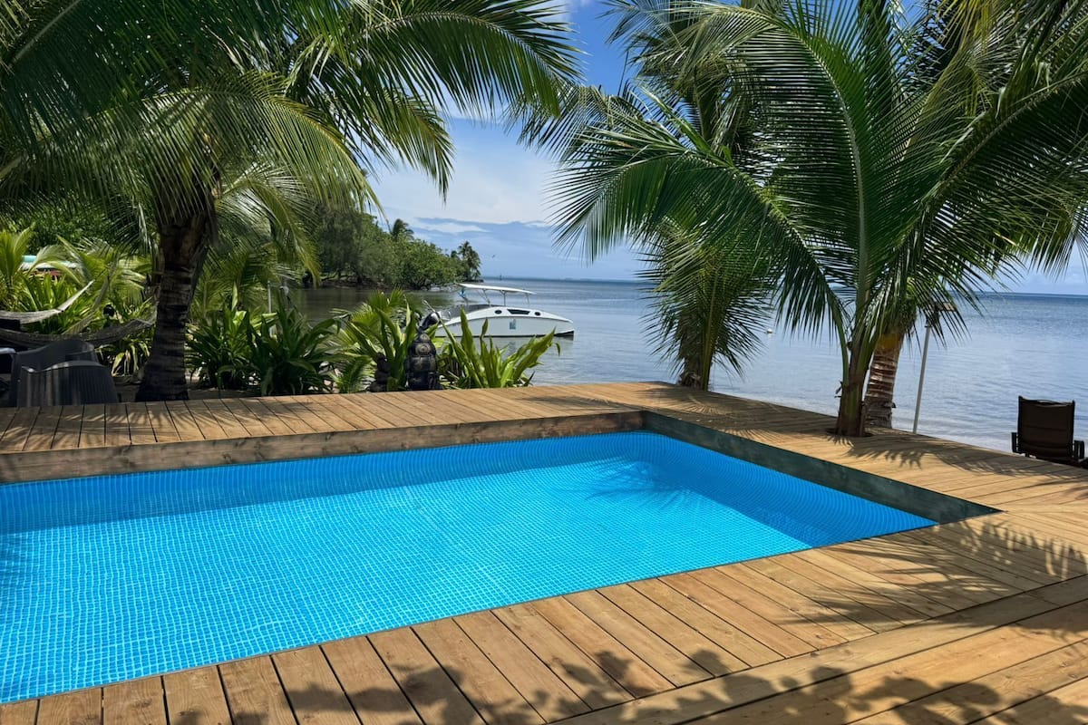 A wooden deck with a clear blue pool, surrounded by palm trees and a view of the ocean.