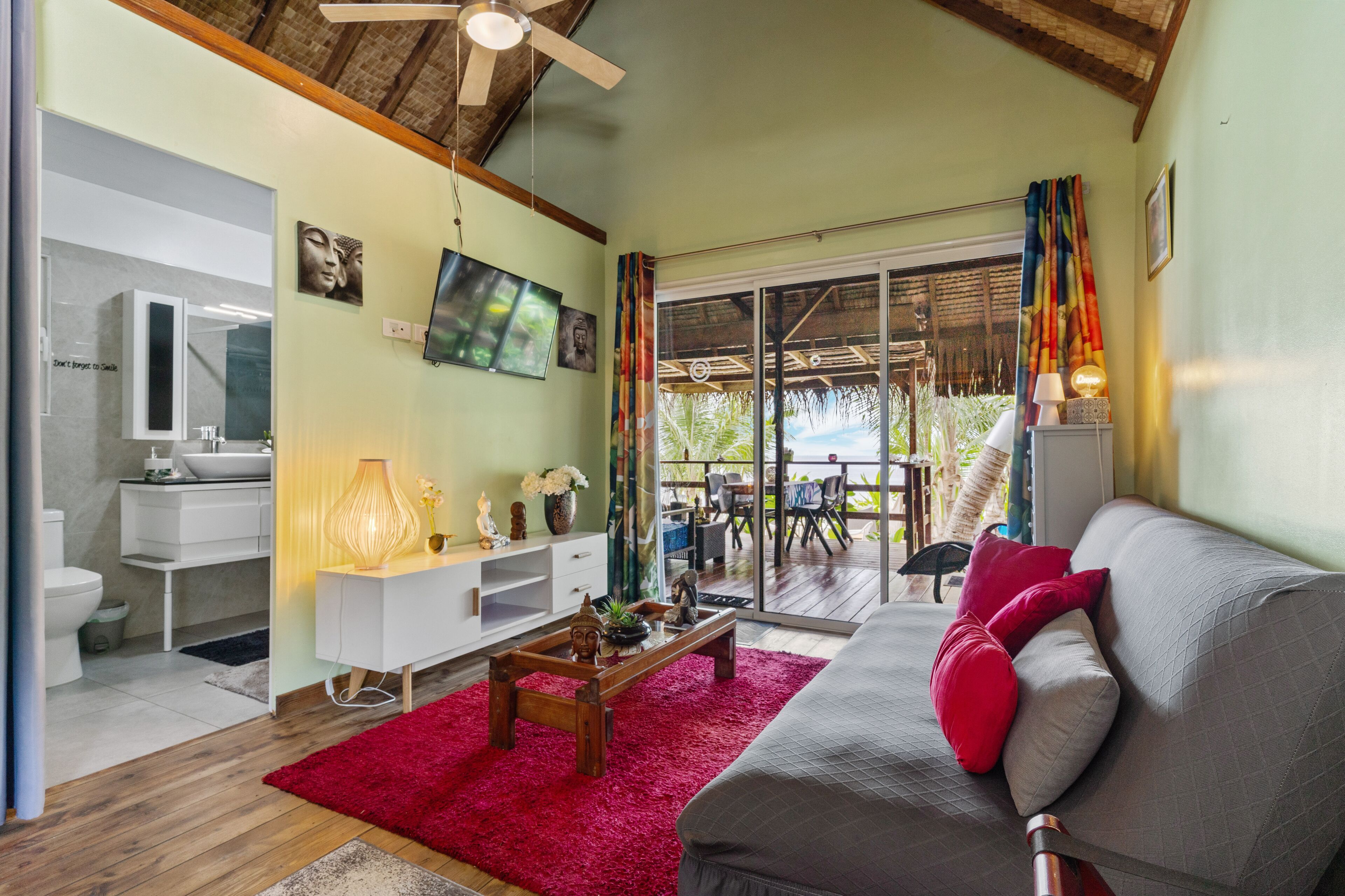 A modern living room with a sofa, a coffee table, and a TV. There is a balcony with a view of greenery.
