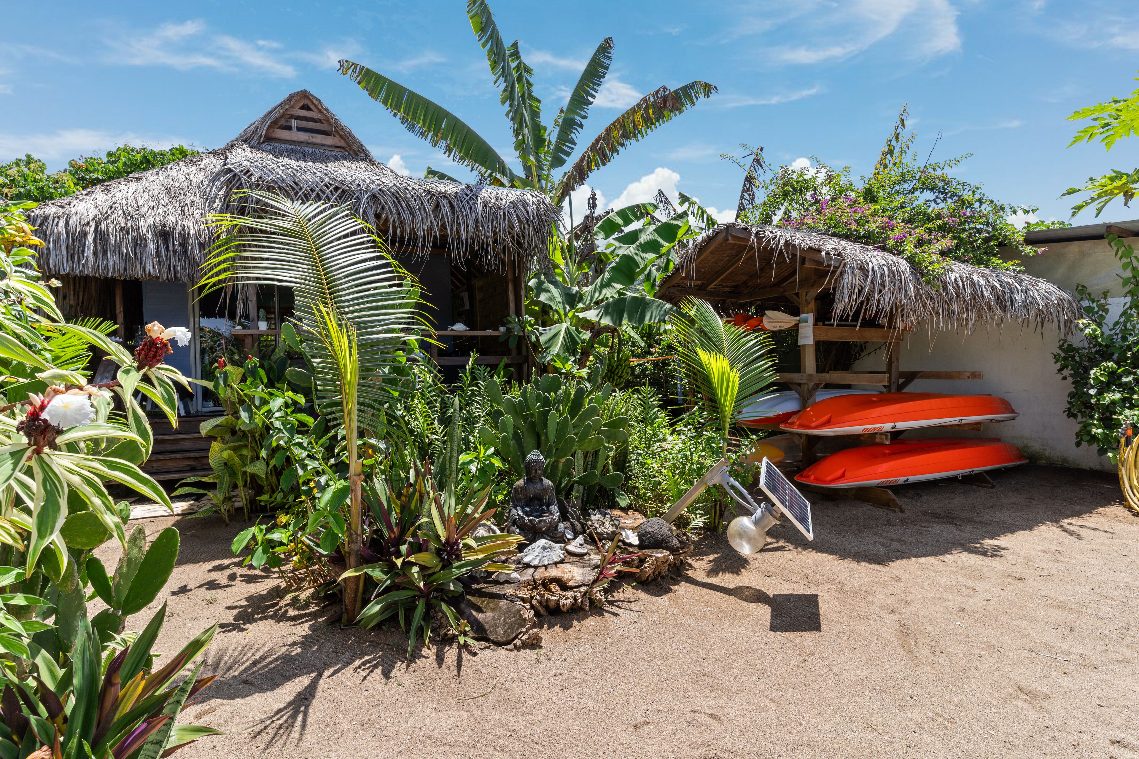 A tropical garden with thatched-roof huts, a statue, and a sandy ground.